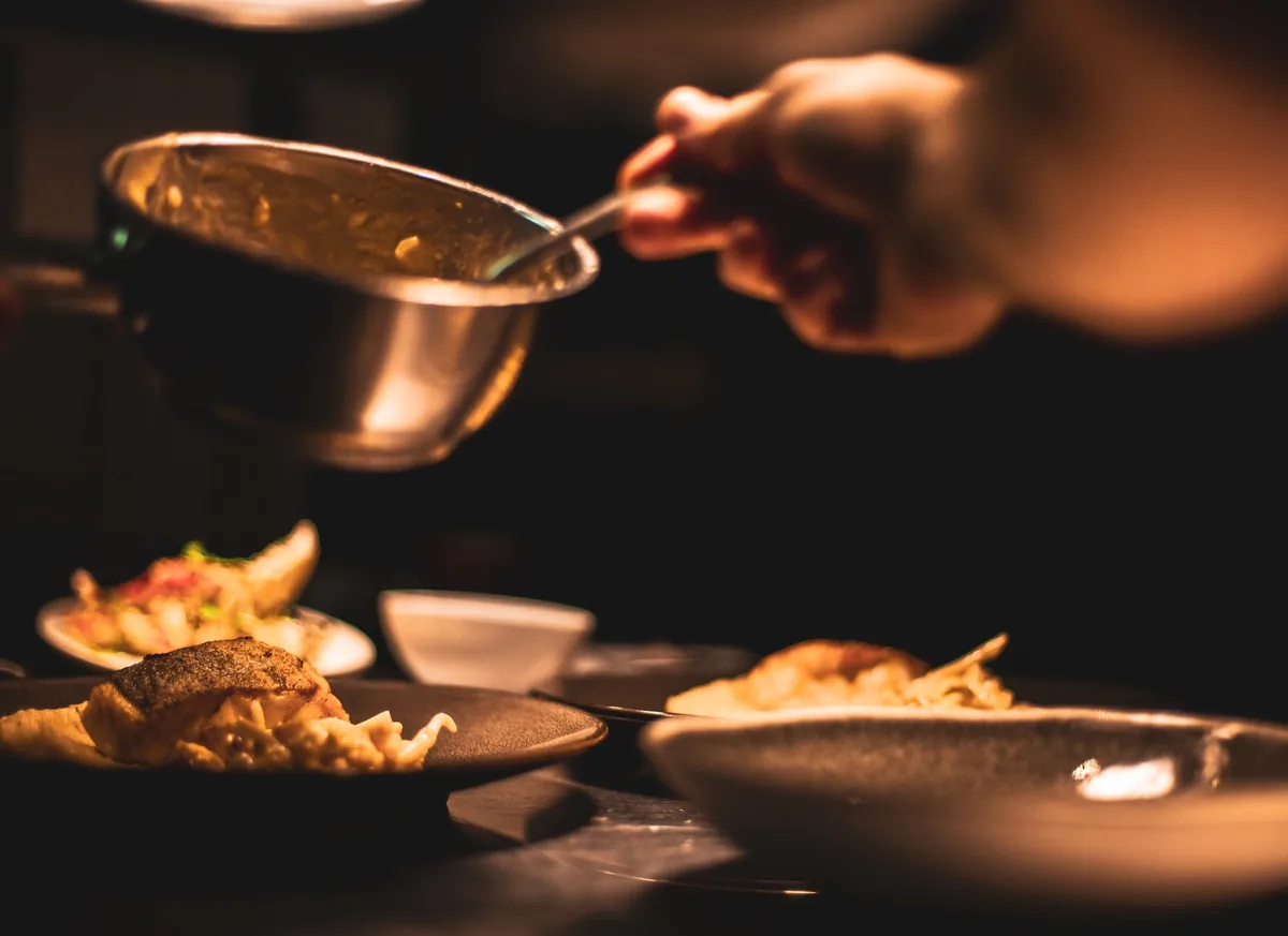 A cook spooning sauce over plates under a warm lamp